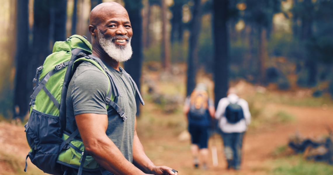 An African American man with a gray hair wearing a green backpack and gray shirt hiking in the woods on a trail.