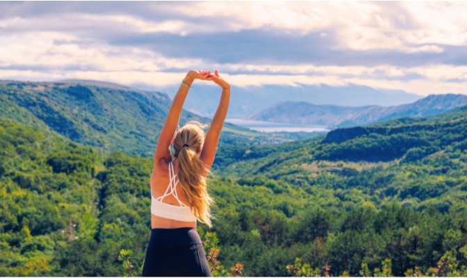 A woman in a white workout top and black leggings stretching with mountains in front of her.