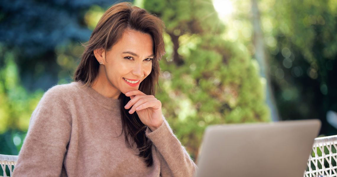 A middle-aged woman with brown hair using her laptop outdoors.