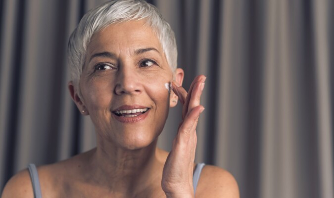 An older woman applying face cream to her face with a gray background.