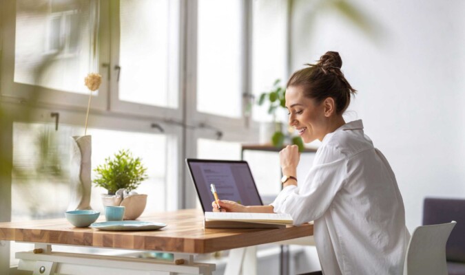 A woman writing at a desk next to a laptop.